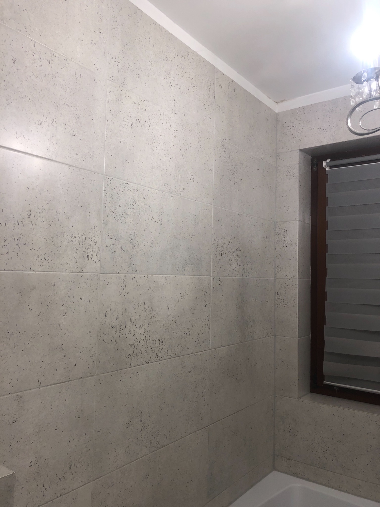 Ściana łazienki wyłożona jasnoszarymi, matowymi płytkami imitującymi kamień z widocznym oknem z roletami w drewnianej ramie i fragmentem wanny.