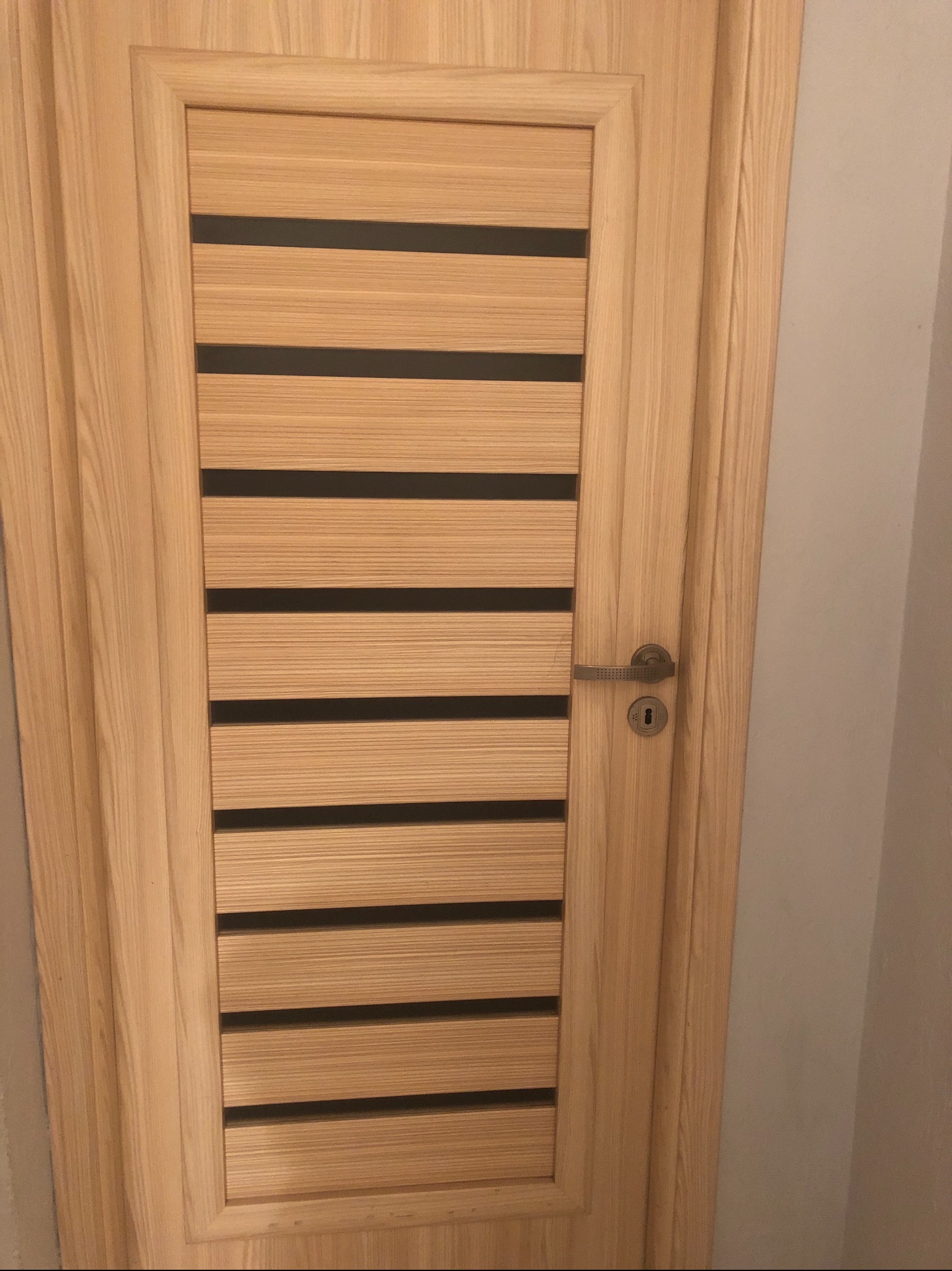Jasne, drewniane drzwi wewnętrzne z poziomymi, czarnymi paskami i srebrną klamką na tle szarej ściany.