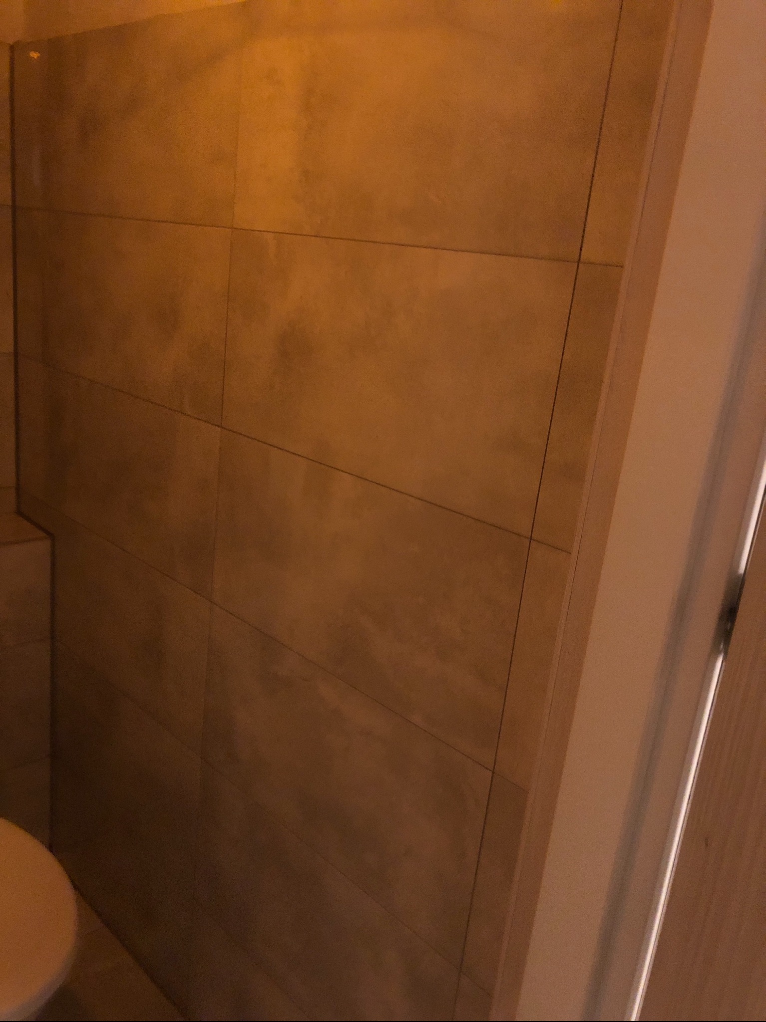 Fragment ściany łazienki wyłożonej brązowymi, matowymi płytkami ceramicznymi, widoczny fragment toalety i ościeżnica drzwi