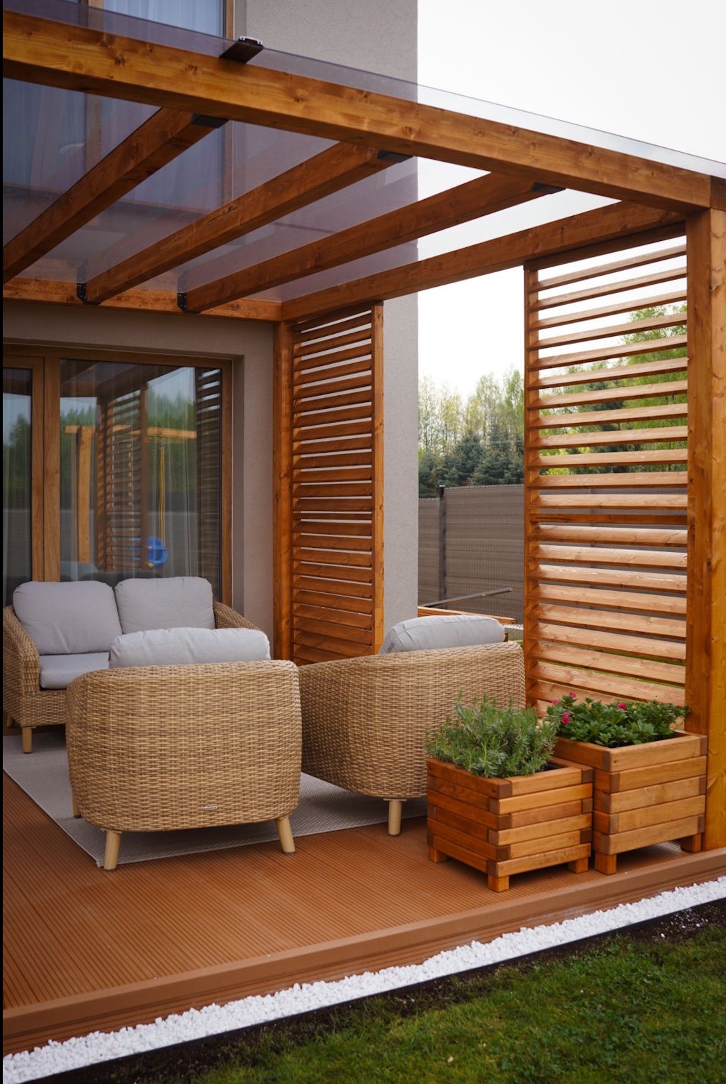 Zadaszona konstrukcja z drewnianą pergolą i lamelami, z brązową podłogą, rattanowymi fotelami i drewnianymi donicami z roślinami.