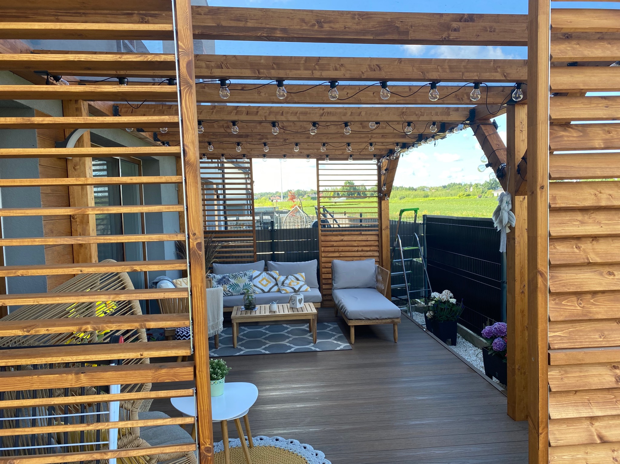 Drewniana pergola z oświetleniem w postaci girlandy żarówkowej, wyposażona w meble ogrodowe, tworząca zaciszne miejsce wypoczynku w ogrodzie w Warszawie.