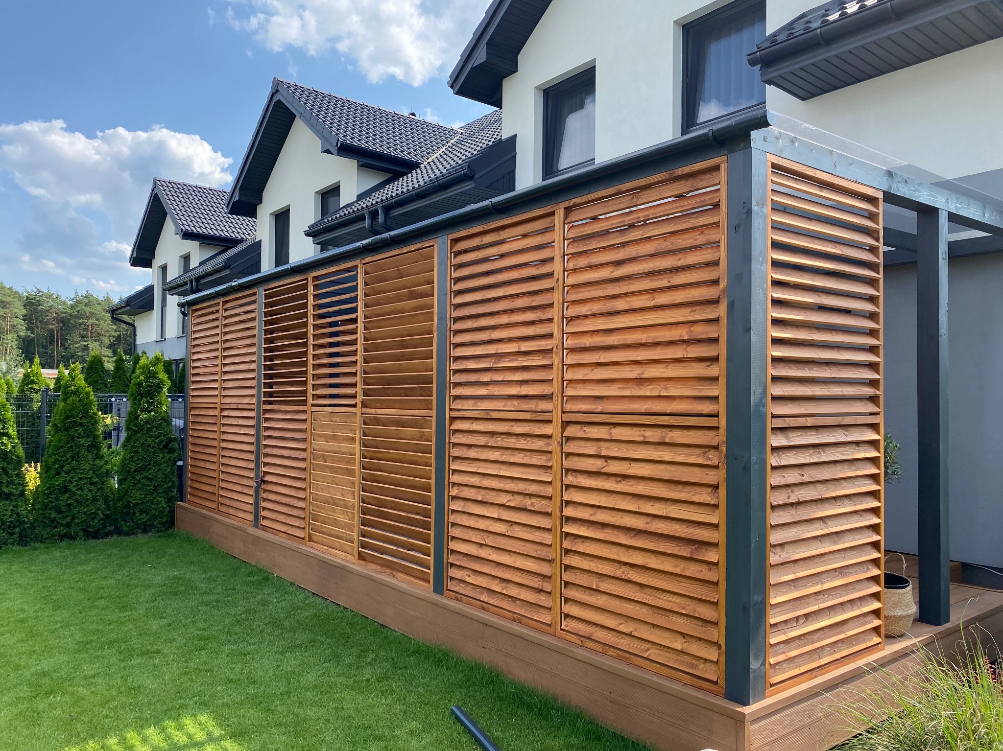 Drewniana konstrukcja z poziomych listew, tworząca ażurową ścianę przy tarasie, z widocznymi elementami konstrukcyjnymi w kolorze grafitowym, na tle zadbanego trawnika i nowoczesnego domu.