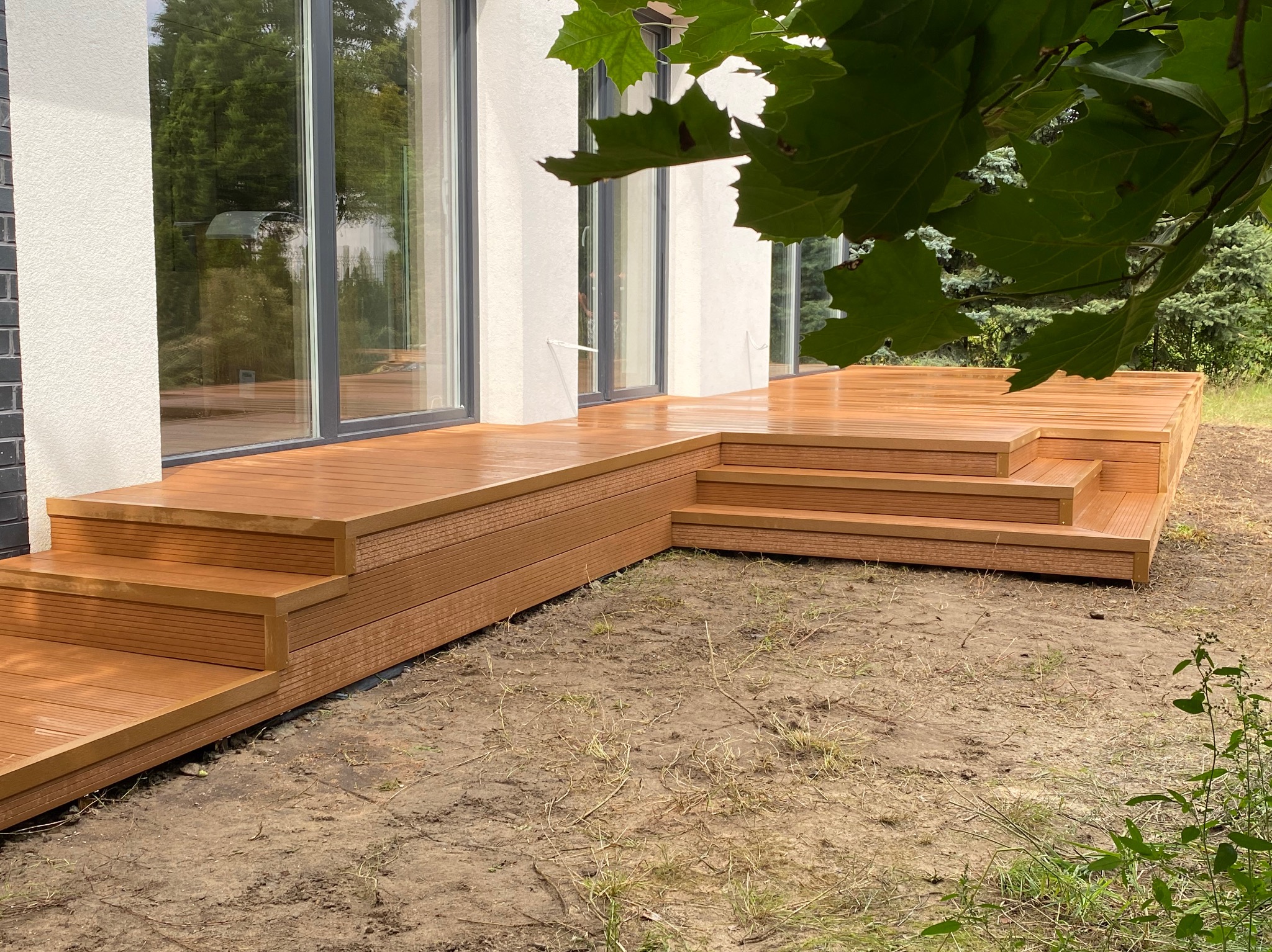 Nowoczesny taras z kompozytu w odcieniach brązu, z niskimi schodami, przylegający do białego domu z dużymi oknami przesuwnymi, widok z poziomu gruntu, z fragmentem zielonych liści w górnej części...