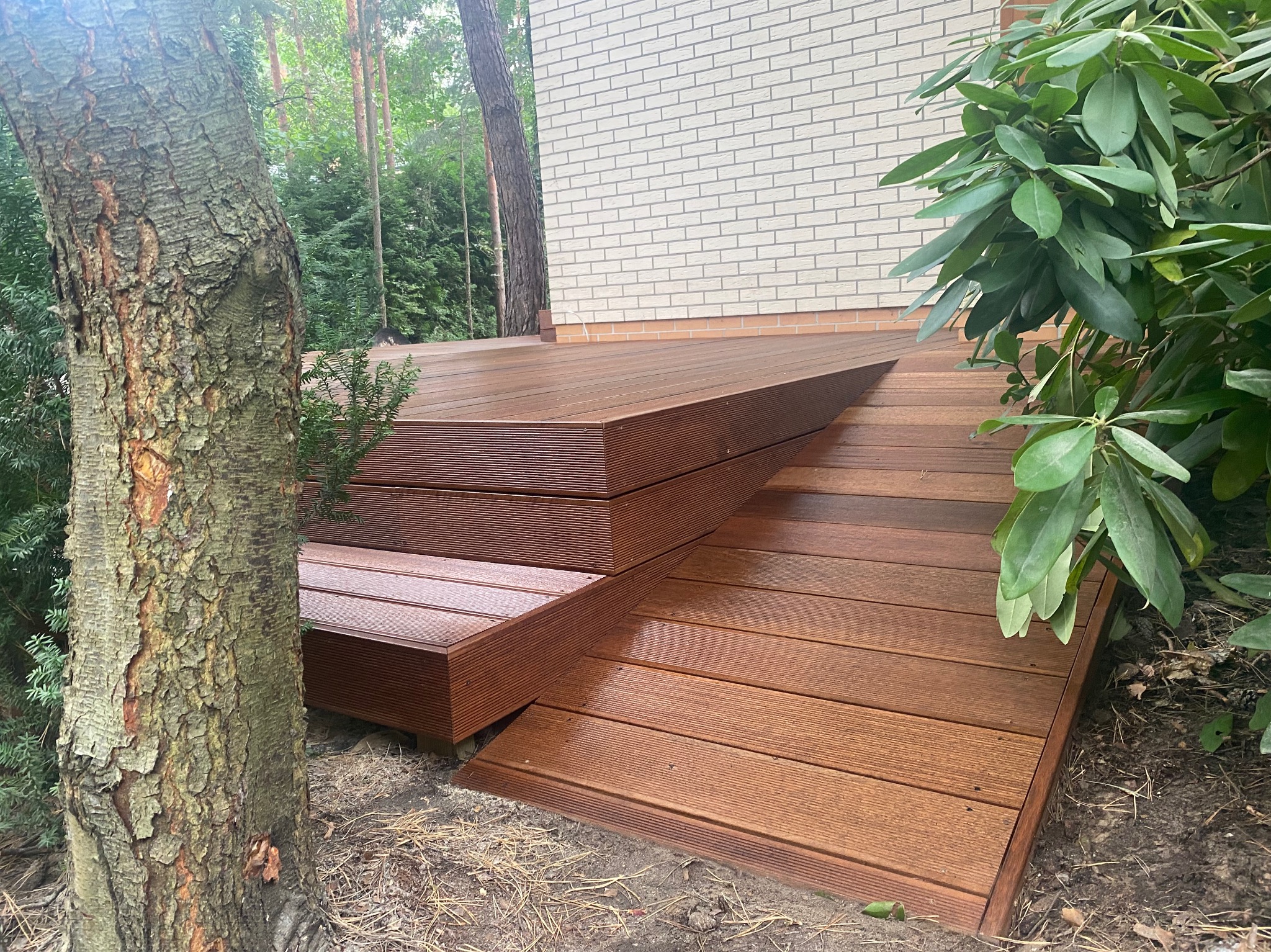 Brązowy taras kompozytowy z wyraźnym usłojeniem, schodami i roślinnością w tle, przylegający do jasnego budynku z cegły.
