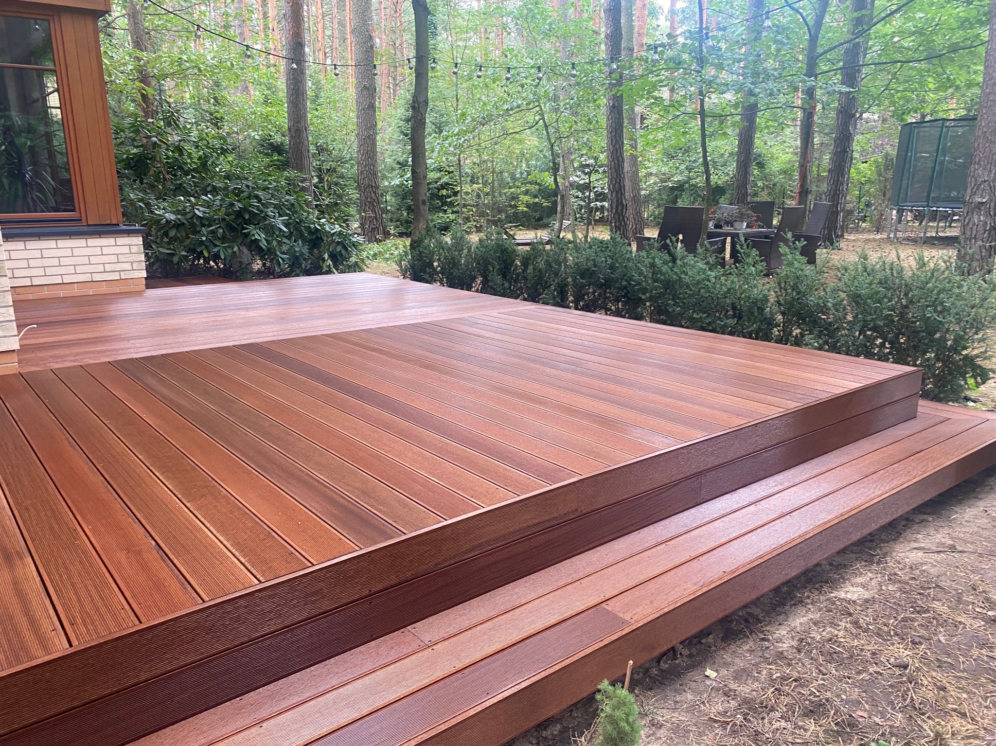 Nowoczesny taras z desek kompozytowych w odcieniach brązu, otoczony zielenią drzew i krzewów, z widocznymi schodami i fragmentem elewacji domu.