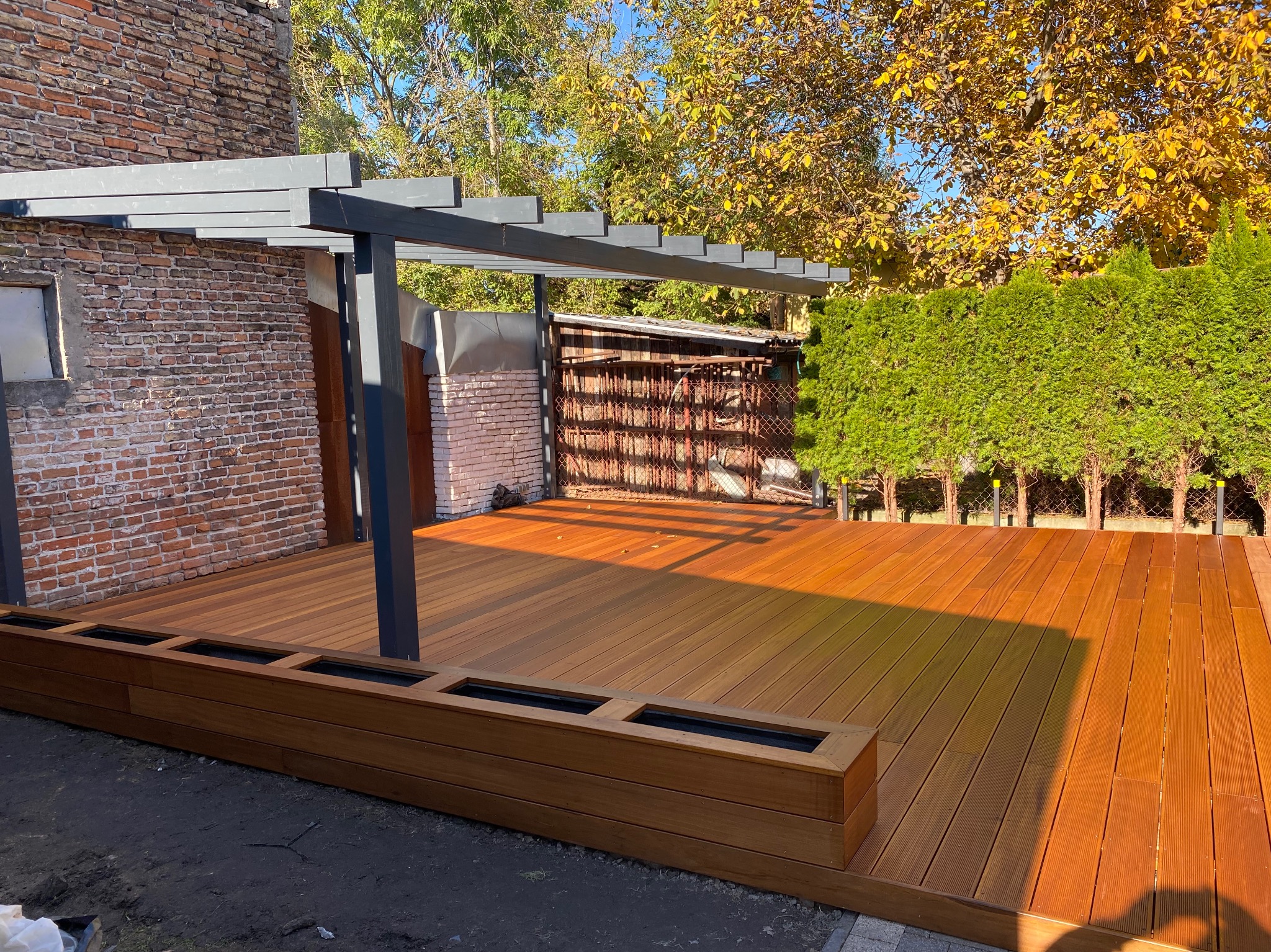 Nowoczesny taras z desek kompozytowych w odcieniach brązu, z szarą pergolą, obok ceglanego budynku i zielonych tuj.