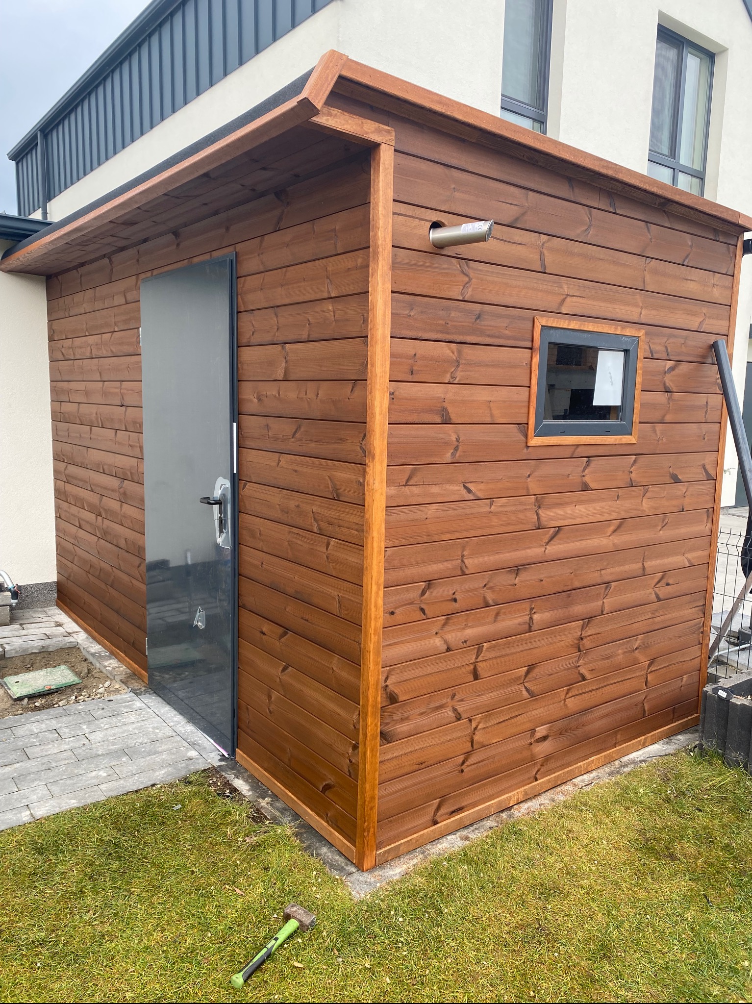 Drewniana konstrukcja z szarą, metalową ramą drzwi i małym oknem, z rurą kominową na zewnątrz, stojąca na trawie obok domu, z młotkiem leżącym na trawniku.