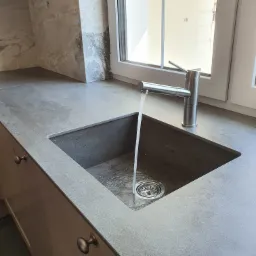 Szary blat kuchenny z wpuszczonym zlewem, bateria z której leci woda, widok na okno i marmurowe wykończenie ściany.