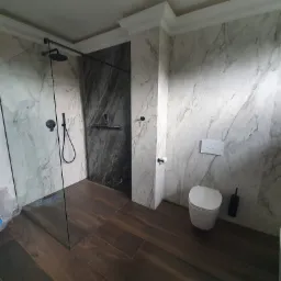 Nowoczesna łazienka z kabiną prysznicową typu walk-in, marmurowymi płytami na ścianach i drewnopodobną podłogą, minimalistyczna toaleta wisząca.