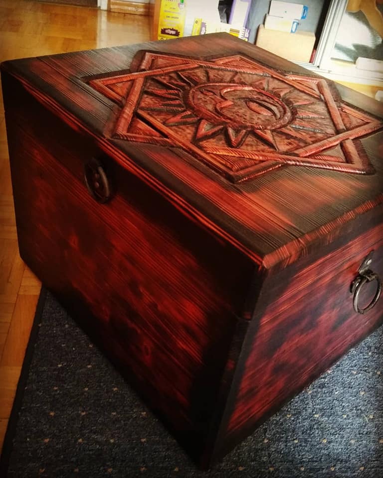 Ręcznie wykonana, drewniana skrzynia w kolorze ciemnej wiśni z rzeźbionym motywem słońca na wieku, widoczne metalowe okucia i tekstura drewna.
