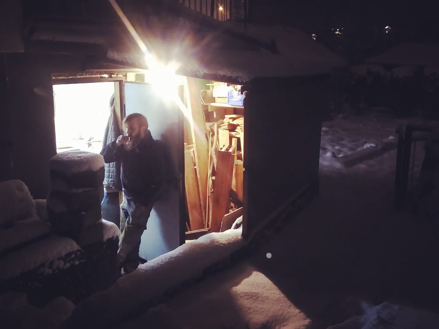 Mężczyzna z brodą pije kawę w wejściu do warsztatu stolarskiego w zimowy wieczór, w tle stos desek i narzędzi.