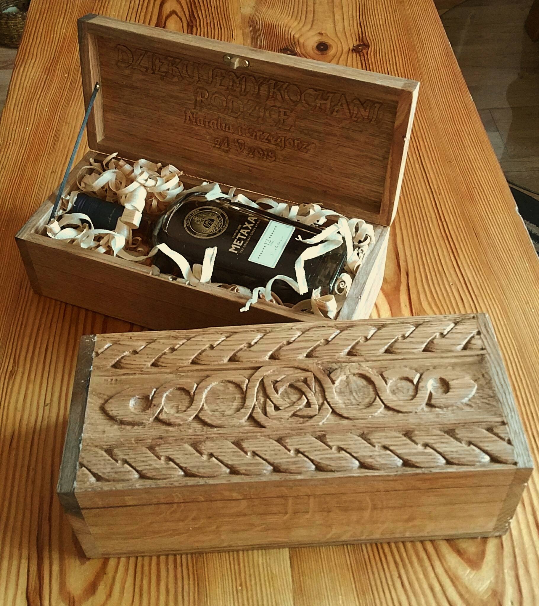 Drewniana skrzynka z wygrawerowanym tekstem 'Dziękujemy Kochanym Rodzicom Natalia i Grzegorz 24.9.2018', wypełniona wiórkami i butelką alkoholu Metaxa, obok druga skrzynka z rzeźbionym wzorem...