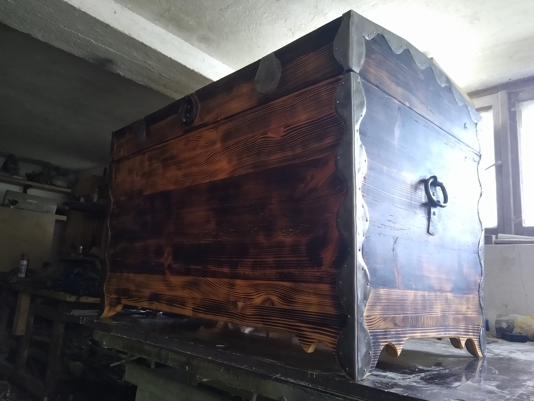 Opalizowana drewniana skrzynia z metalowymi okuciami, widoczna w warsztacie, stoi na stole roboczym pod betonowym stropem.