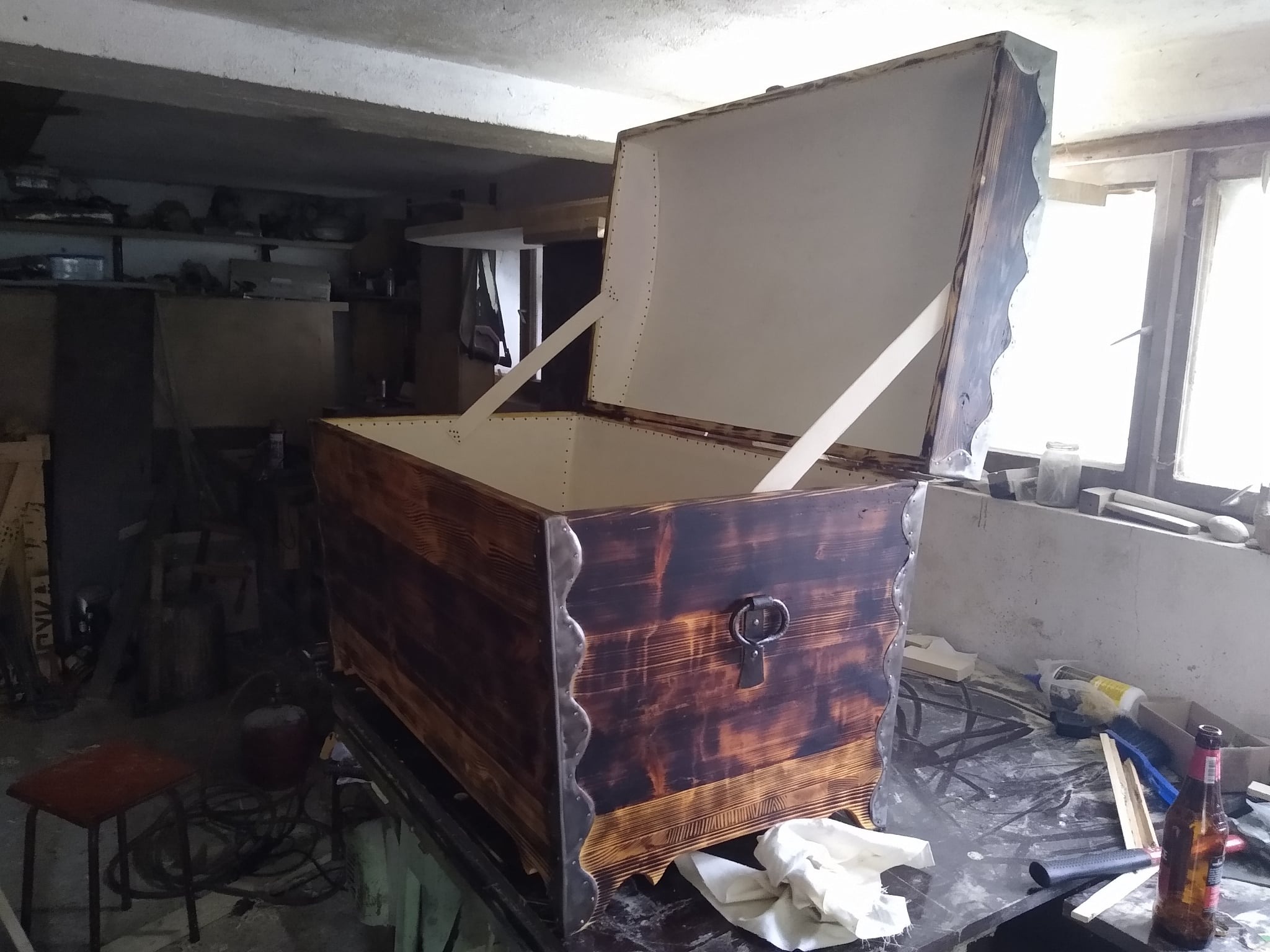 Otwarta, ręcznie robiona skrzynia z ciemnego, wypalanego drewna z jasnym, pomalowanym wnętrzem, widoczne metalowe zdobienia i robocze otoczenie.