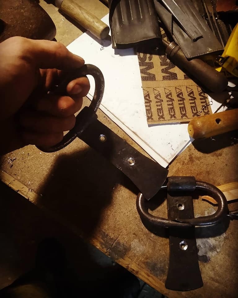 Ręcznie kute, metalowe okucia z uchwytami, prawdopodobnie do drzwi, w trakcie obróbki na warsztacie stolarskim, widoczne narzędzia i materiały.