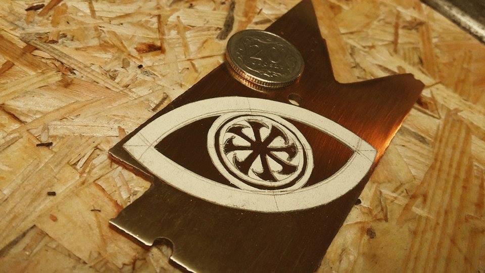Miedziany wycięty kształt oka z narysowanym wzorem i monetą na wierzchu, leżący na płycie OSB.