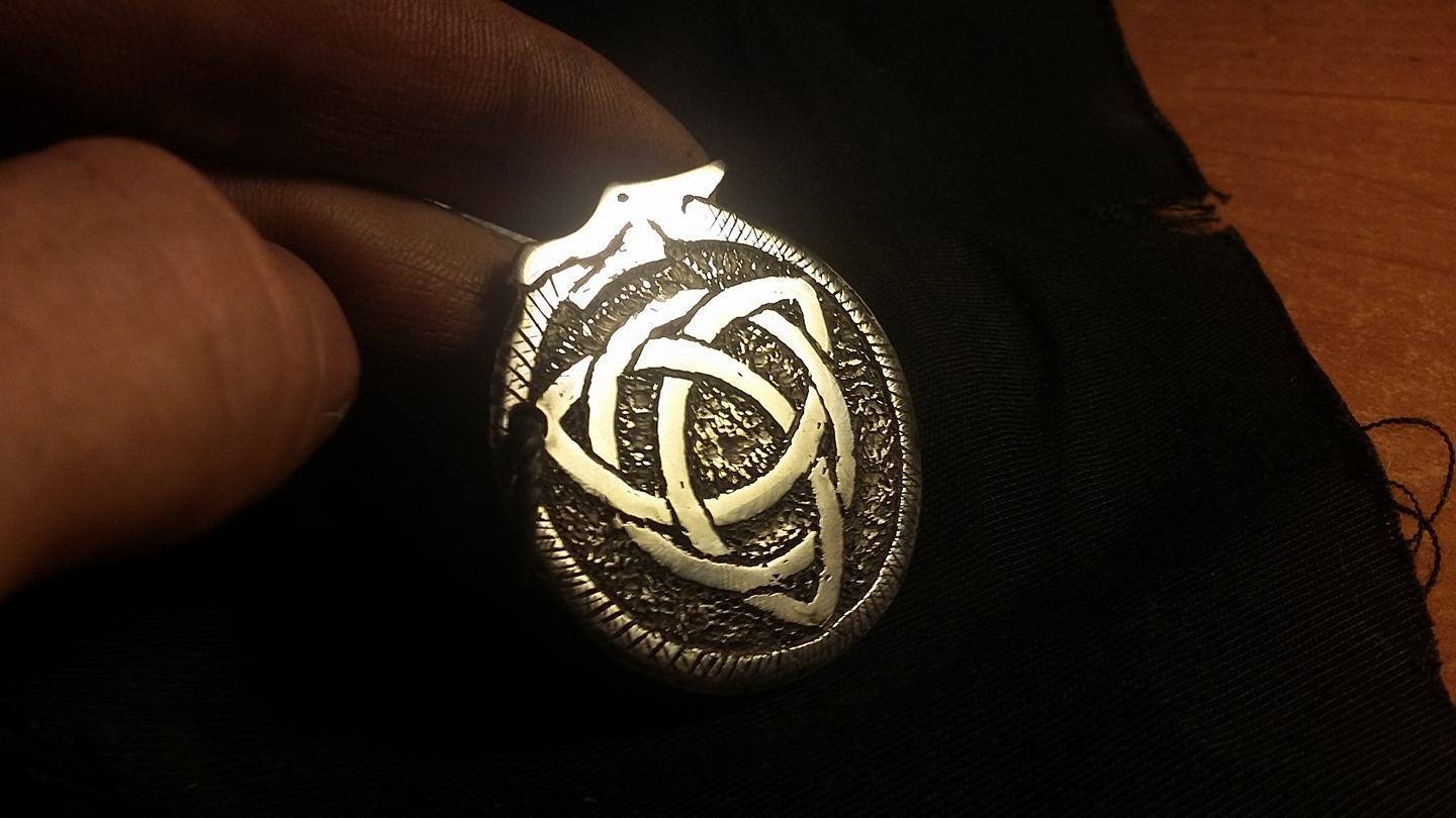 Owalny, srebrny wisiorek z celtyckim węzłem trójkątnym, trzymany w palcach na tle czarnej tkaniny i drewnianej powierzchni.