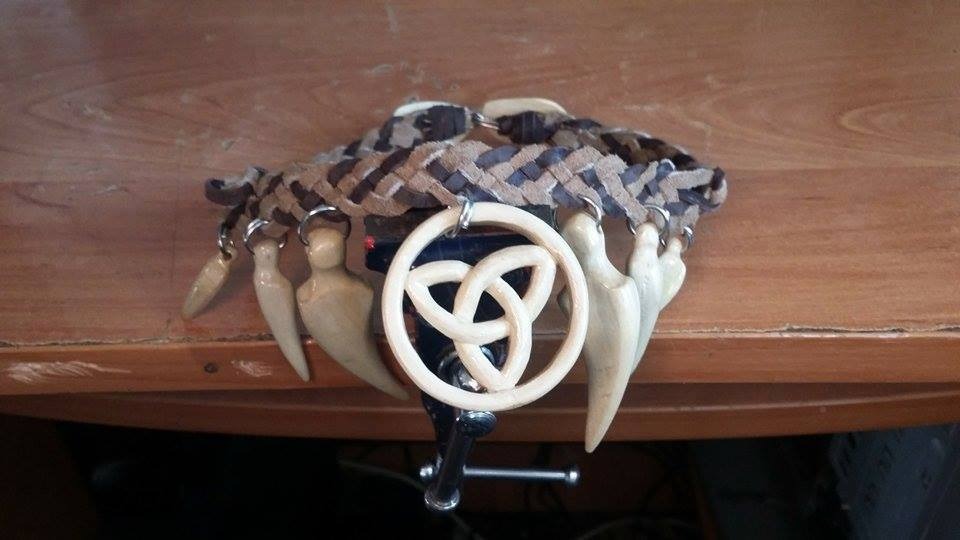 Ręcznie robiony naszyjnik z plecionego sznurka, zawieszkami w kształcie kłów i centralnym elementem z wyżłobionym symbolem celtyckim, leżący na drewnianym stole.