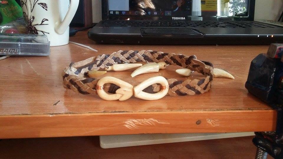 Ręcznie robiony, pleciony z brązowej i beżowej skóry naszyjnik z drewnianymi elementami i wstawkami z zębów leżący na drewnianym biurku z laptopem i kubkiem w tle.