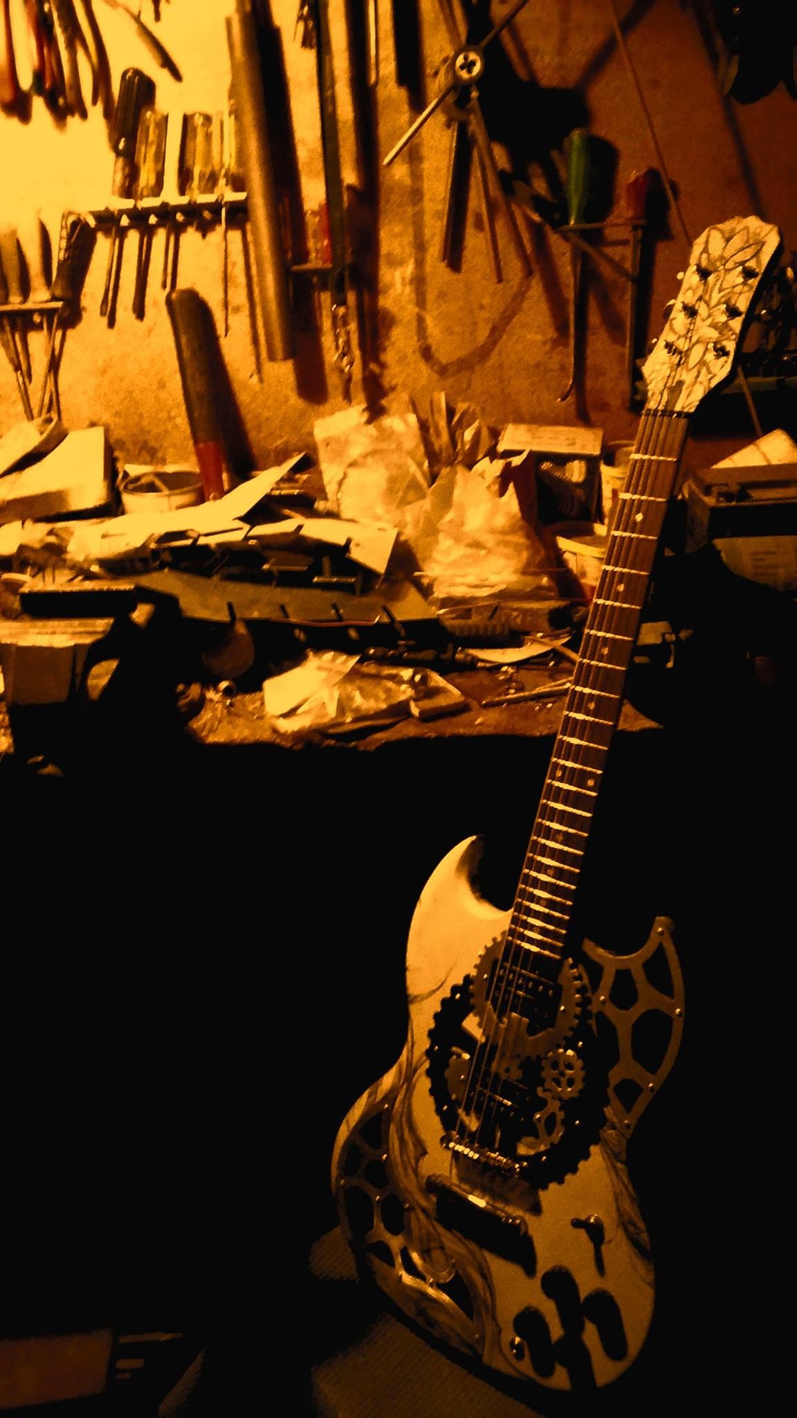 Niestandardowa gitara elektryczna z motywem steampunk, z widocznymi zębatkami i wzorami na korpusie, stojąca w warsztacie z narzędziami na ścianie i bałaganem na stole.