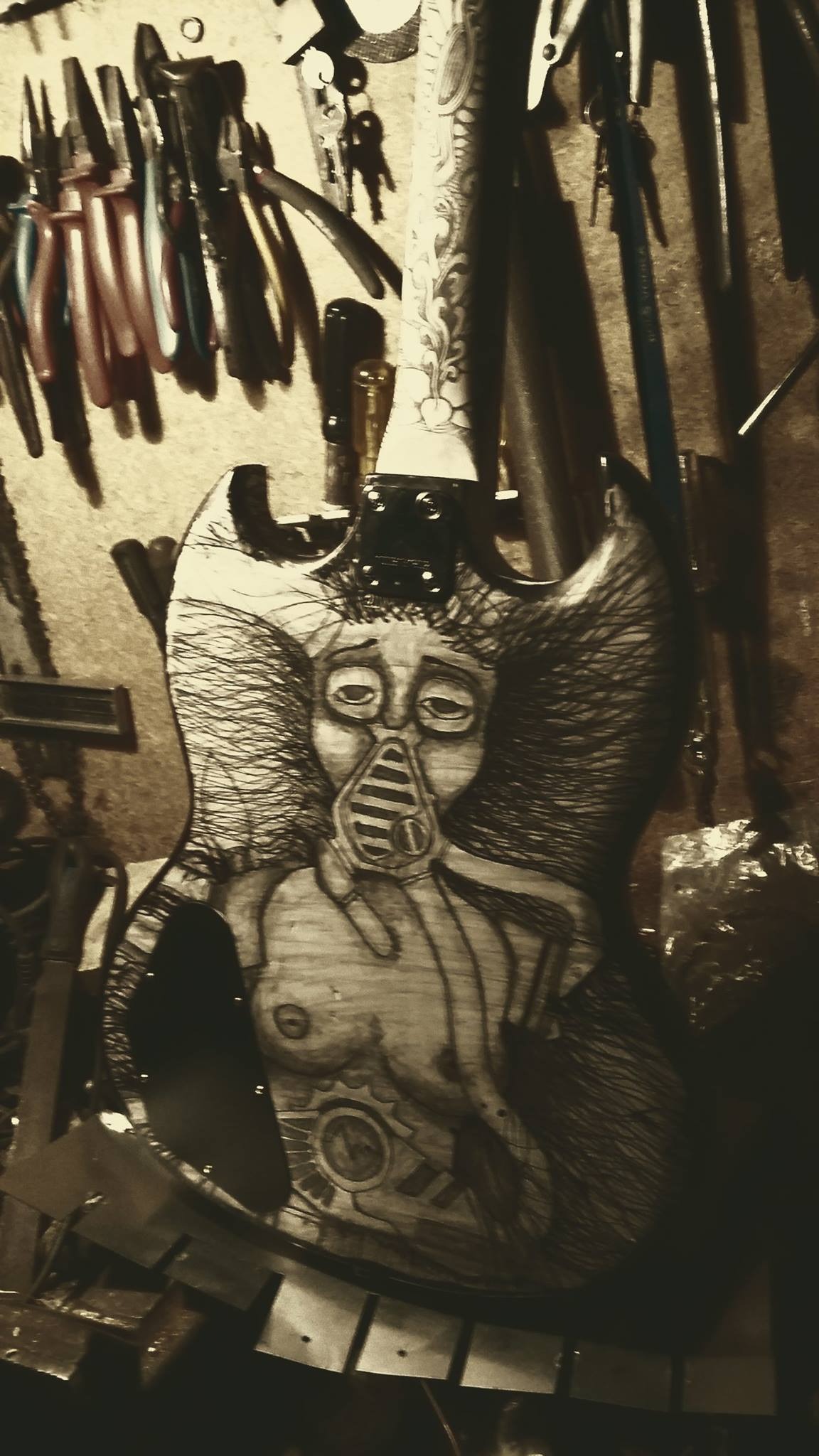 Gitara elektryczna z ręcznie malowanym, mrocznym motywem postaci w masce gazowej, wisząca w warsztacie na tle narzędzi.