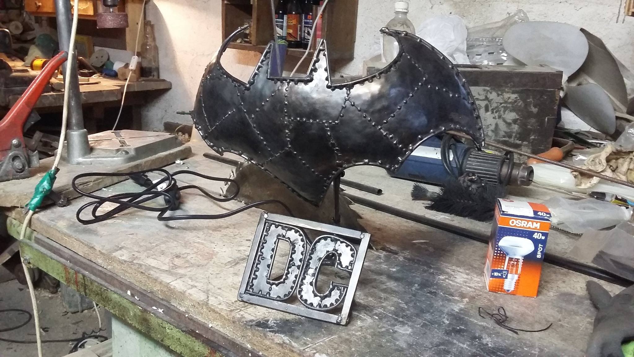 Metalowa rzeźba w kształcie symbolu Batmana i logo DC wykonane z metalowych elementów, stojące na stole warsztatowym w warsztacie, widoczna skrzynka z żarówkami Osram