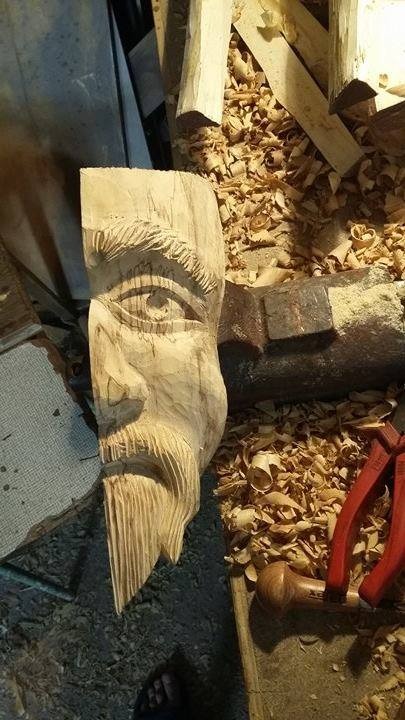 Niedokończona rzeźba w drewnie przedstawiająca profil twarzy mężczyzny z brodą, otoczona wiórami i narzędziami stolarskimi.