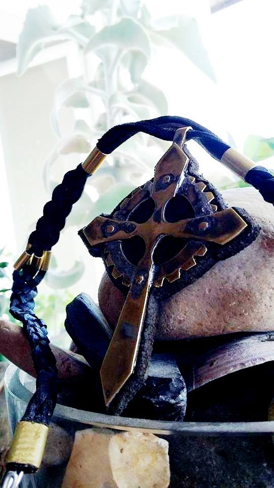 Dekoracyjny krzyż z elementami metalowymi i czarnym sznurkiem z metalowymi zdobieniami, umieszczony na kamieniach w szklanym naczyniu, z rośliną w tle.