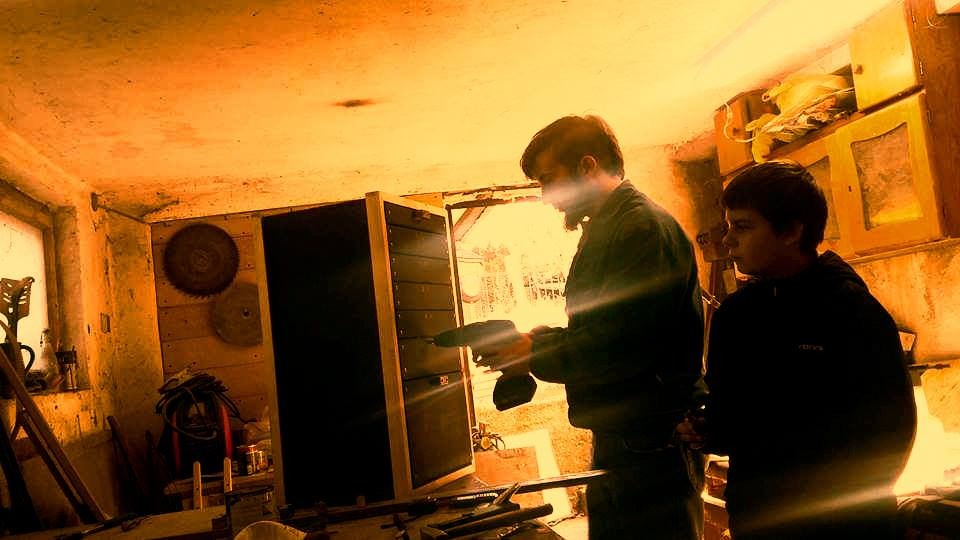 Dwóch mężczyzn w warsztacie, jeden wierci w ciemnych drzwiach z jasną ramą, na ścianie wiszą tarcze do piły, widoczne narzędzia i bałagan.