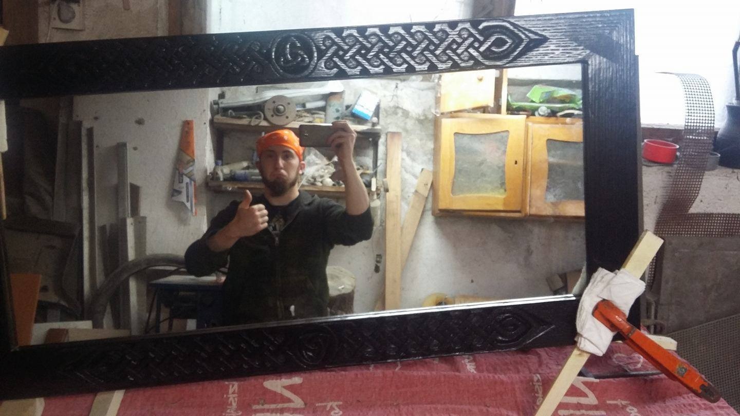 Mężczyzna w pomarańczowej chuście robi selfie w lustrze z ręcznie rzeźbioną, czarną ramą w stylu celtyckim, które stoi na stole warsztatowym z pomocą ścisków stolarskich.