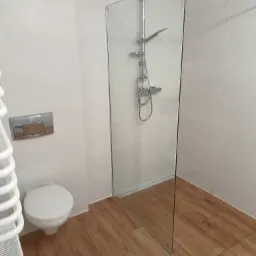 Nowoczesna łazienka z kabiną prysznicową typu walk-in ze szklaną ścianką, białą armaturą i grzejnikiem drabinkowym, wykończona płytkami imitującymi drewno na podłodze i białymi płytkami na ścianach.