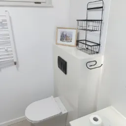 Biała toaleta z zabudowaną spłuczką, nad którą wisi czarna, metalowa półka z koszykami i ramka ze świątecznym obrazkiem. Widoczny również biały grzejnik drabinkowy i rolka papieru toaletowego.