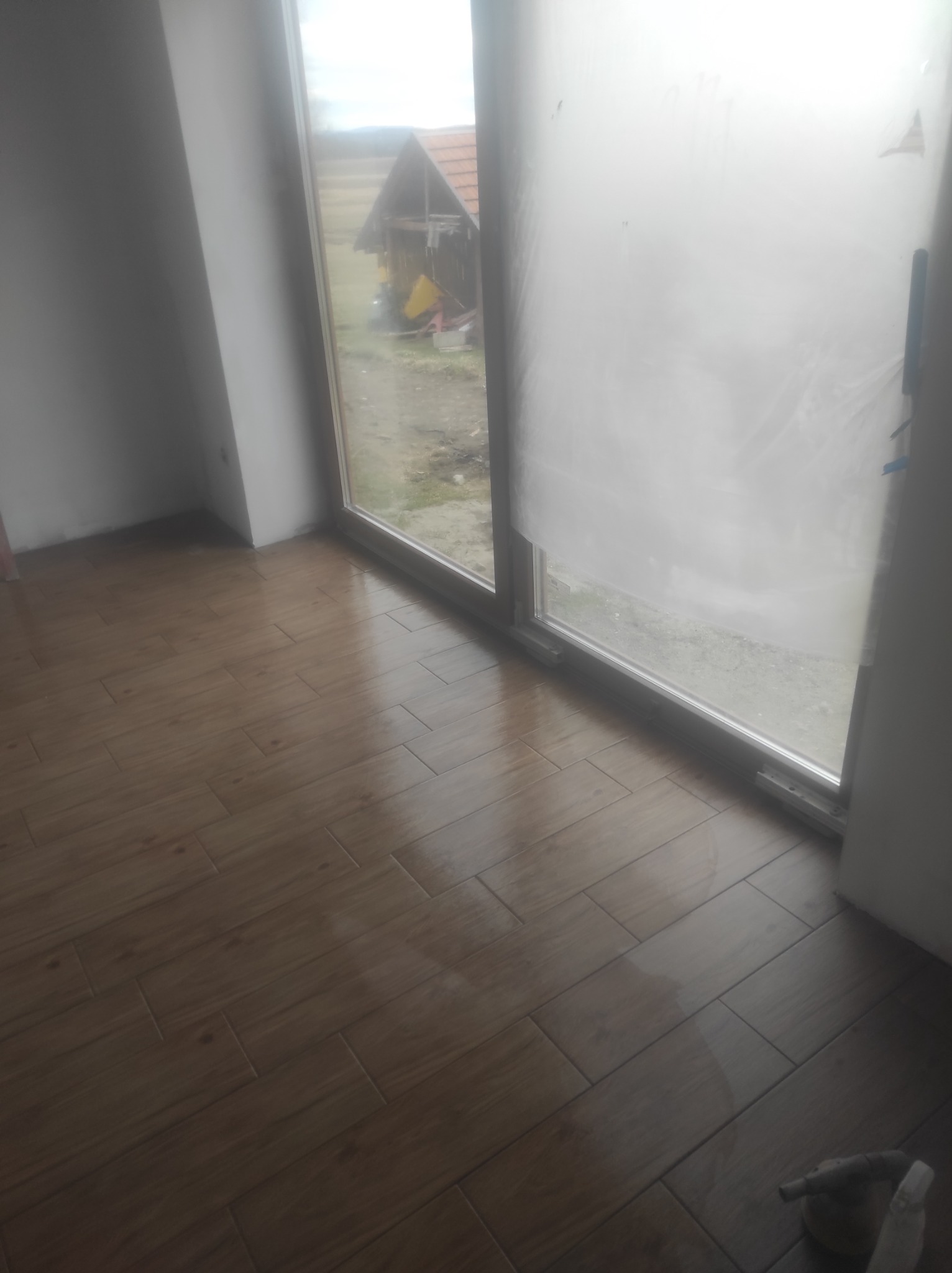 Ułożona podłoga z brązowych płytek imitujących drewno, widok z góry na dużą oszkloną ścianę z widokiem na zewnątrz, częściowo zasłoniętą folią ochronną.