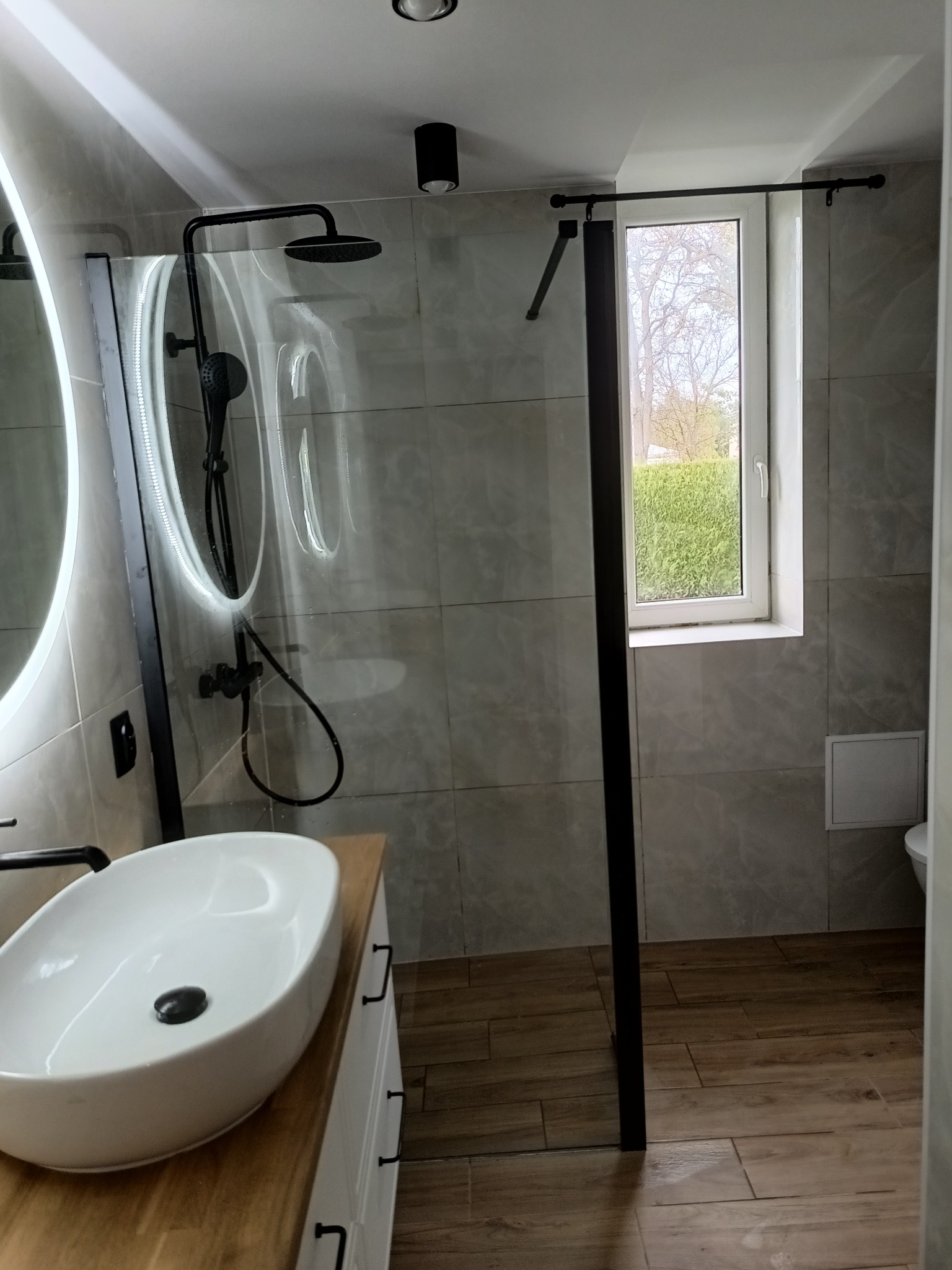 Nowoczesna łazienka z kabiną prysznicową typu walk-in z czarnymi elementami, owalnym lustrem z podświetleniem LED, umywalką nablatową na drewnianym blacie i widokiem z okna na zieleń.
