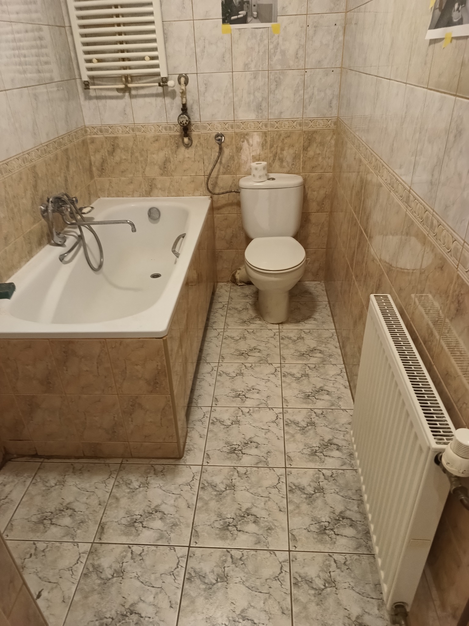 Standardowa łazienka z wanną, toaletą, grzejnikiem drabinkowym i beżowo-szarą glazurą z dekoracyjnym pasem. Na ścianie przyklejone zdjęcia.