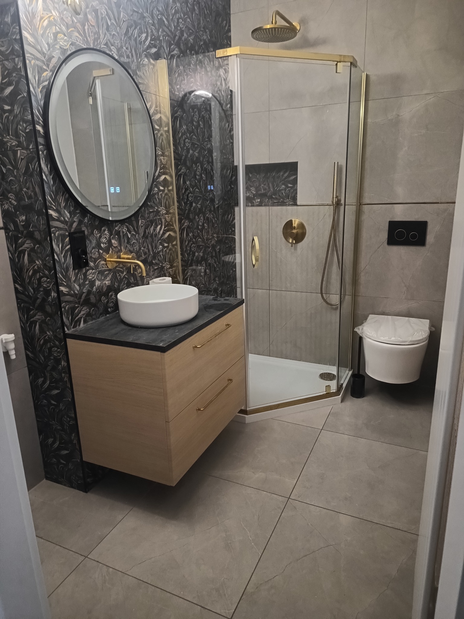 Nowoczesna łazienka po remoncie z kabiną prysznicową ze złotymi detalami, okrągłym lustrem z podświetleniem LED nad drewnianą szafką z umywalką nablatową i toaletą z czarnym przyciskiem spłukującym.