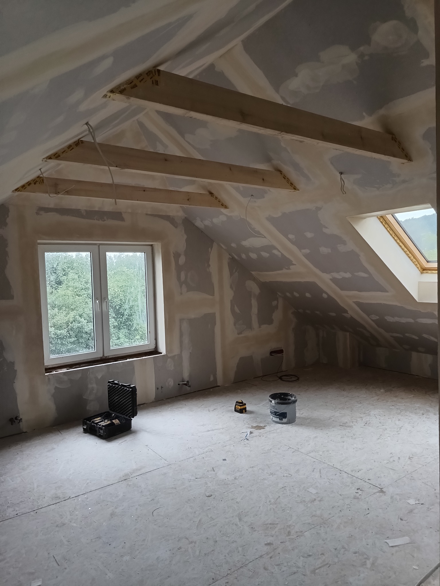 Poddasze w trakcie adaptacji: widoczne drewniane belki konstrukcyjne, okna połaciowe, ściany pokryte płytami gipsowo-kartonowymi w trakcie szpachlowania, narzędzia w kuferku na podłodze.