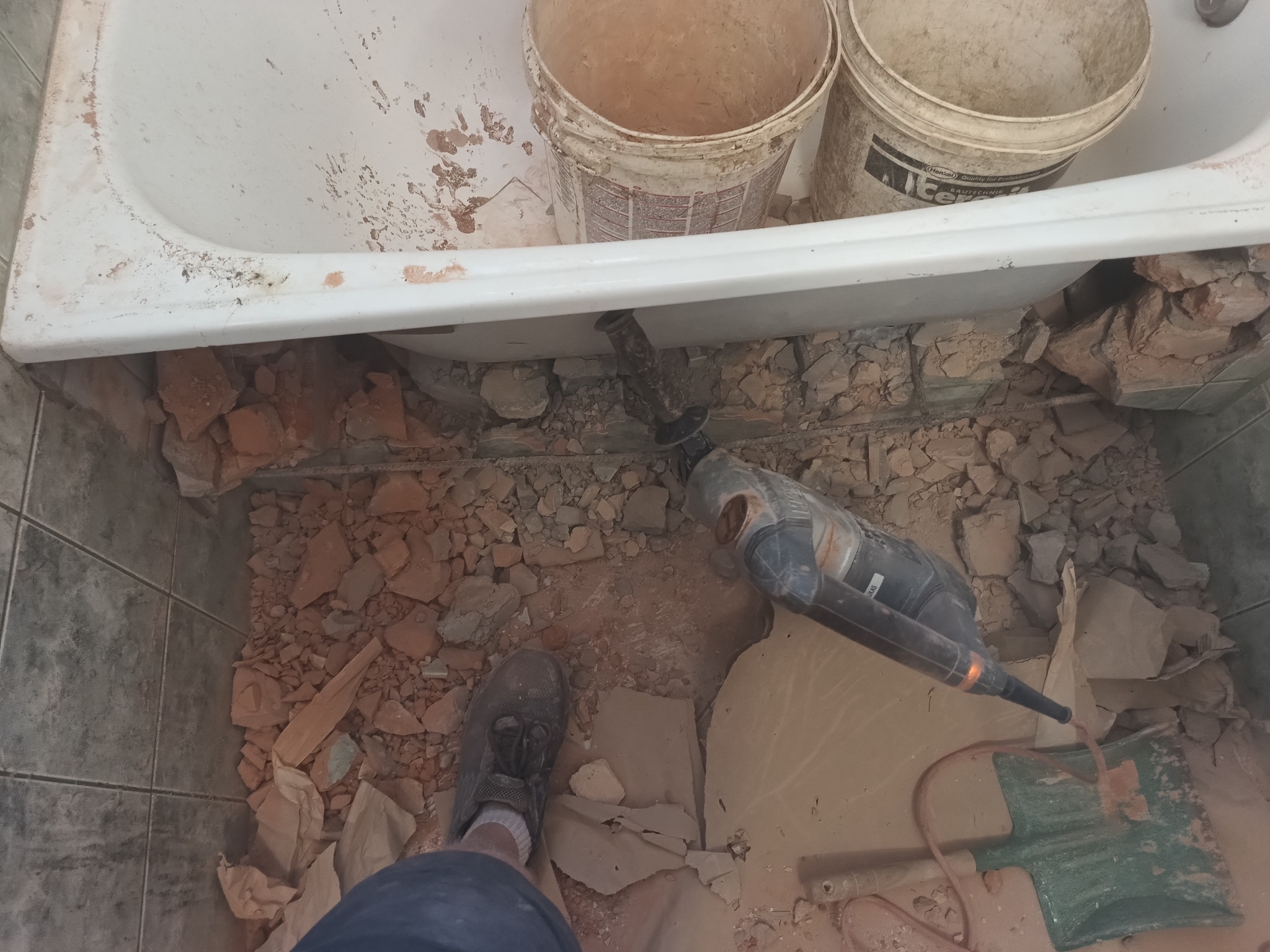 Demontaż płytek pod wanną: widoczny gruz, wiertarka udarowa, szufelka i fragment buta osoby wykonującej prace.