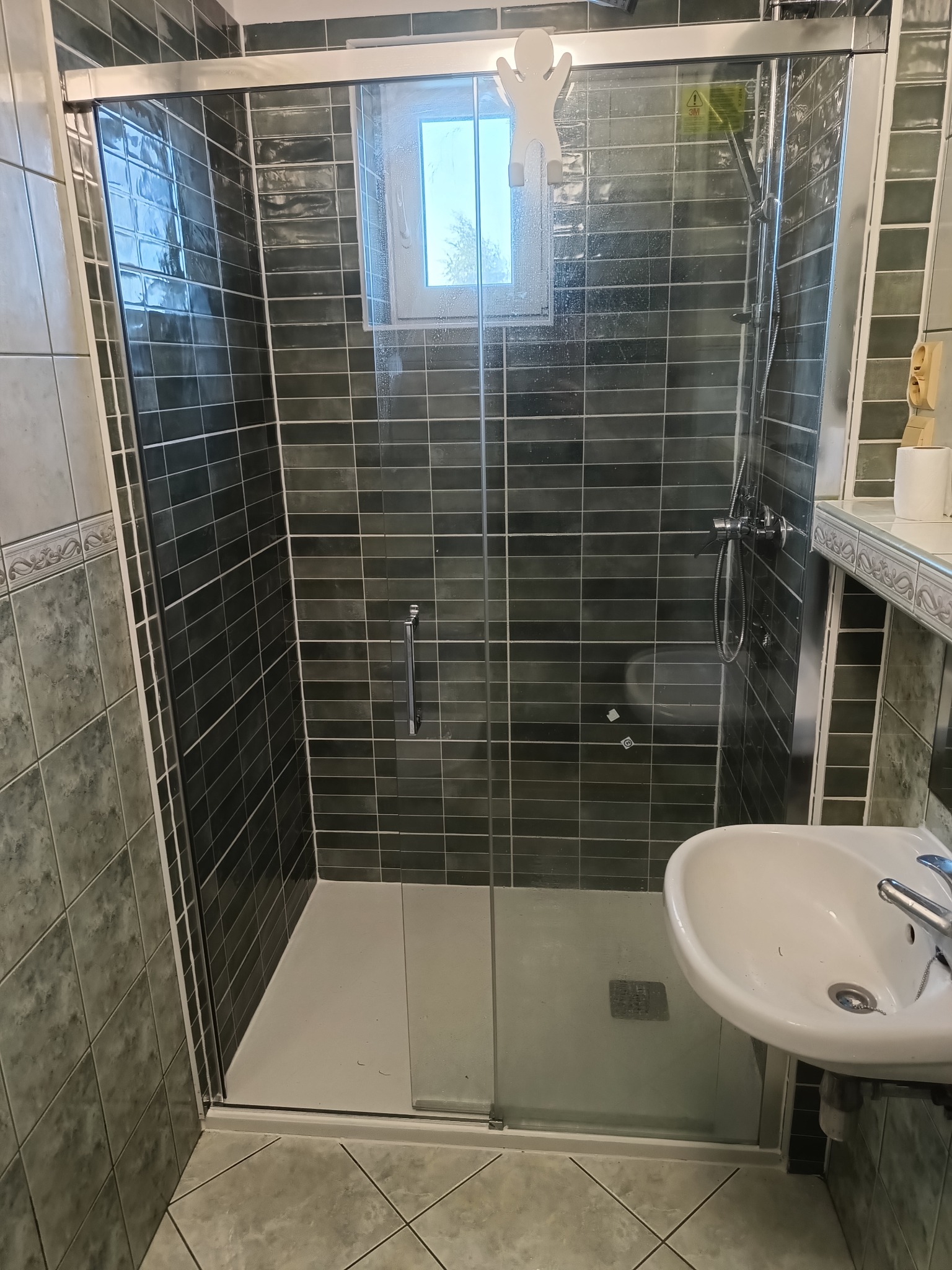 Kabina prysznicowa z przesuwnymi drzwiami, wyłożona ciemnymi, poziomymi płytkami, z widoczną umywalką i oknem z naklejką w kształcie ludzika.