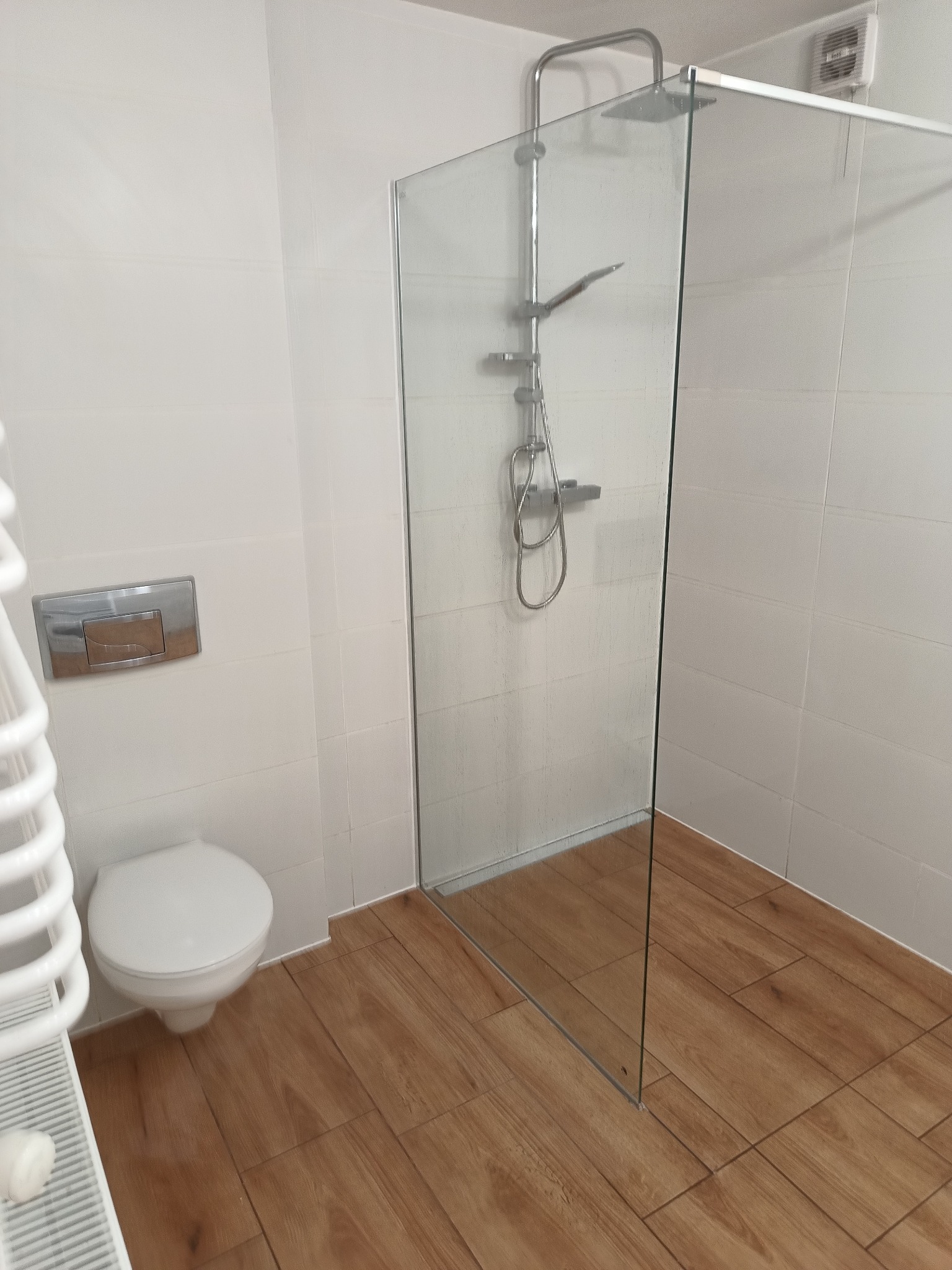 Nowoczesna łazienka z kabiną prysznicową typu walk-in ze szklaną ścianką, białą armaturą i grzejnikiem drabinkowym, wykończona płytkami imitującymi drewno na podłodze i białymi płytkami na ścianach.