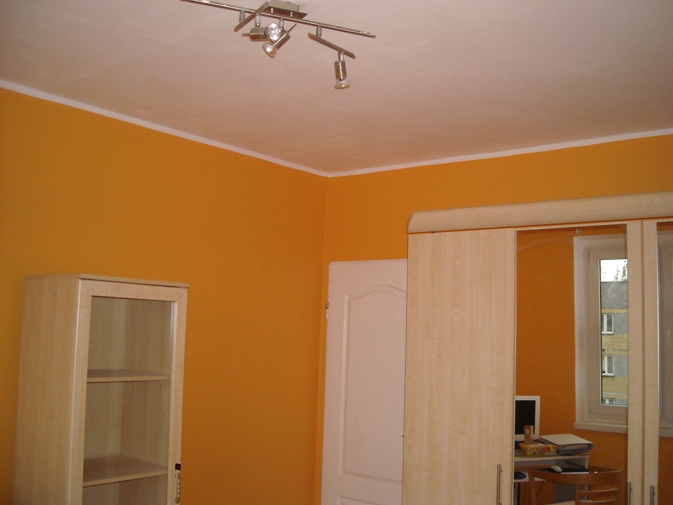 Pomarańczowa ściana po malowaniu z widoczną gładzią, biały sufit z reflektorami, meble w kolorze jasnego drewna.