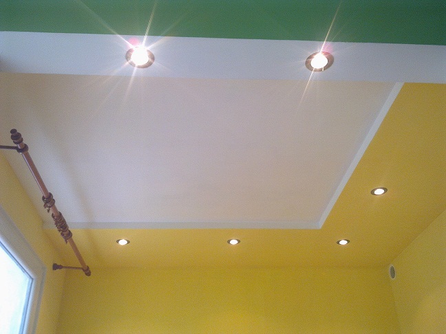 Pomarańczowo-żółte ściany i biały sufit podwieszany z oświetleniem punktowym, widoczny fragment karnisza przy oknie.