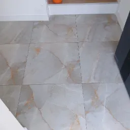 Wykończona podłoga z płytek imitujących marmur z pomarańczowym żyłkowaniem, widoczny fragment schodów z drewnopodobnymi panelami i pomarańczowe wiadro z narzędziami glazurniczymi.