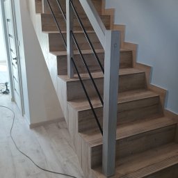 Remont wykaczenia wnętrz - Wykończone schody z drewnopodobnych płytek z czarnymi metalowymi balustradami i jasnymi drewnianymi poręczami, widoczne narzędzia i kable w tle.