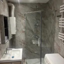 Wykończona łazienka z kabiną prysznicową, umywalką z szafką, pralką i grzejnikiem drabinkowym na ścianie z płytkami imitującymi marmur.