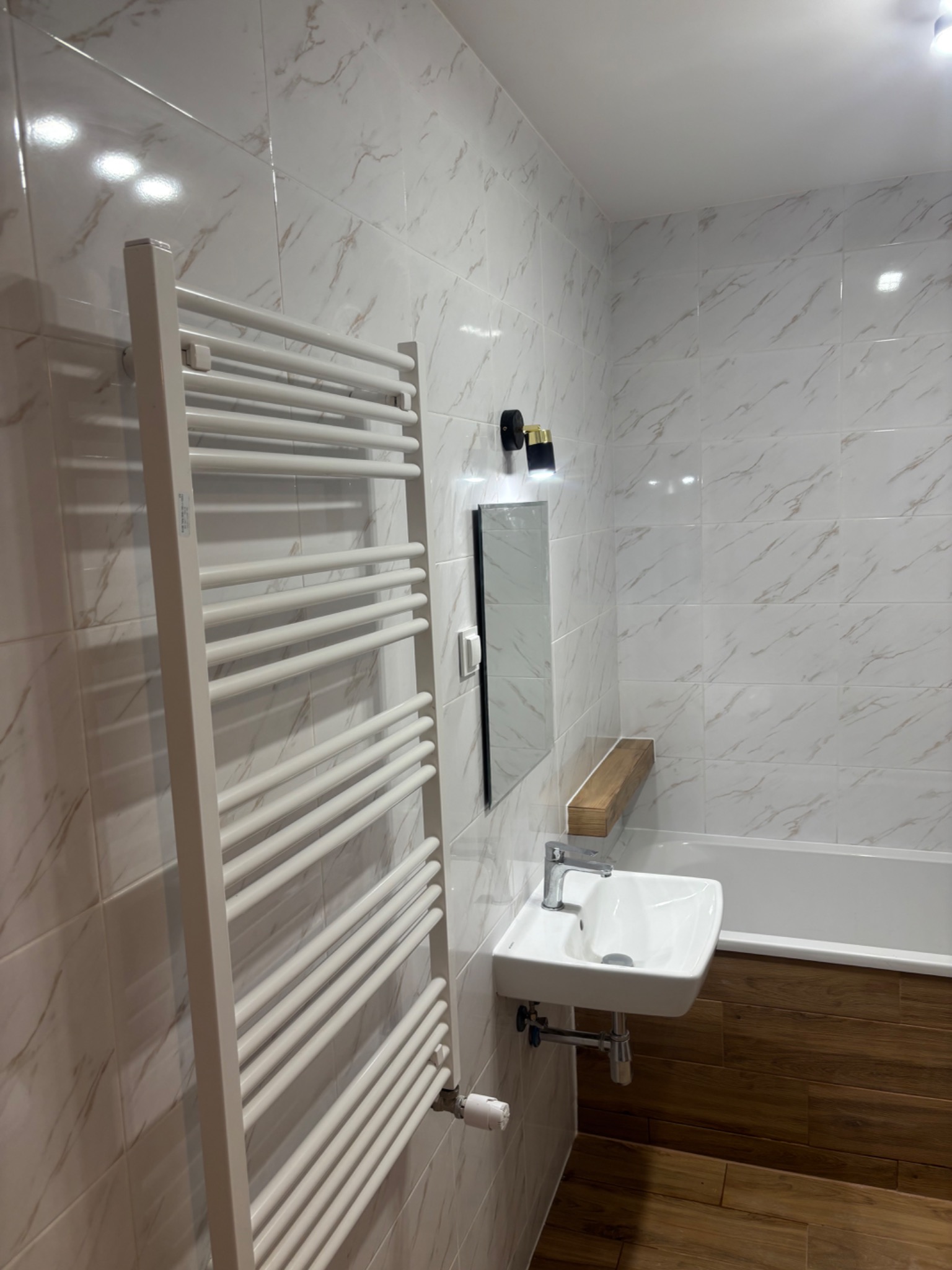 Wykończona łazienka z białymi marmurkowymi płytkami, grzejnikiem drabinkowym, umywalką nablatową i drewnianymi akcentami.