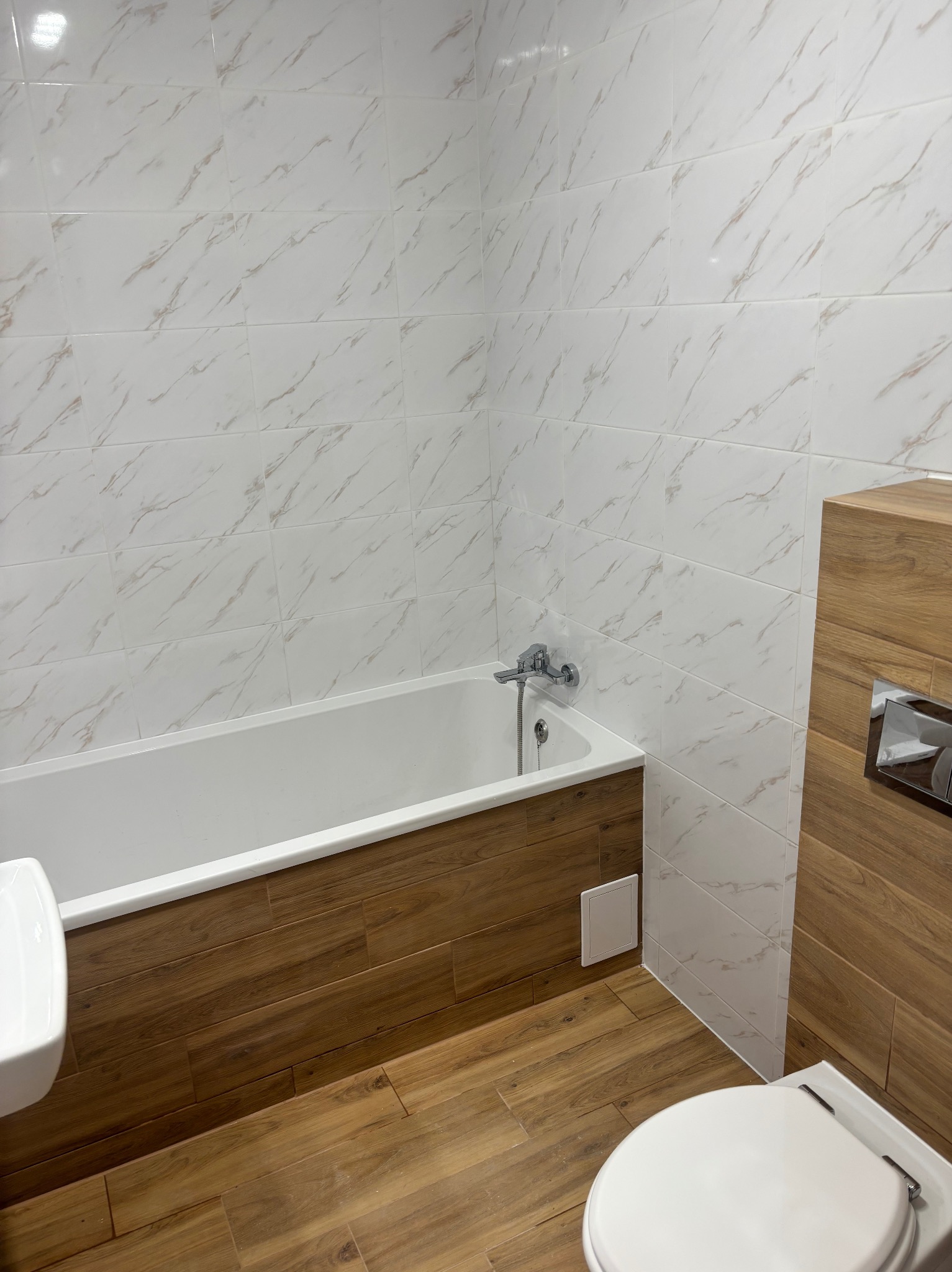 Wykończona łazienka z wanną obudowaną drewnopodobnymi panelami, białe płytki imitujące marmur na ścianach i drewnopodobna podłoga, widoczna toaleta i umywalka.