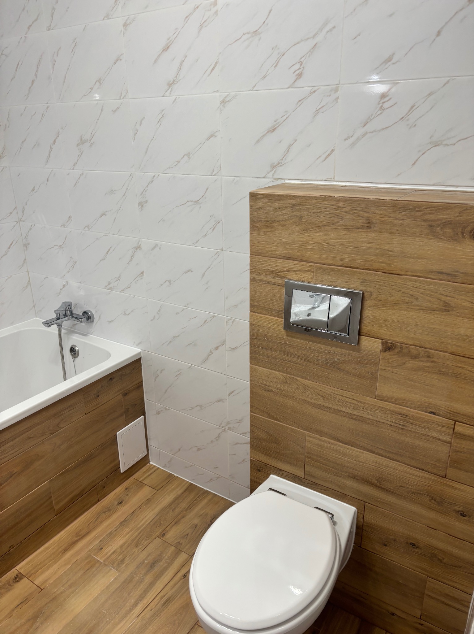 Nowoczesna łazienka z białymi płytkami imitującymi marmur na ścianach, drewnopodobnymi płytkami na podłodze i zabudowie WC, chromowana bateria wannowa i przycisk spłuczki.
