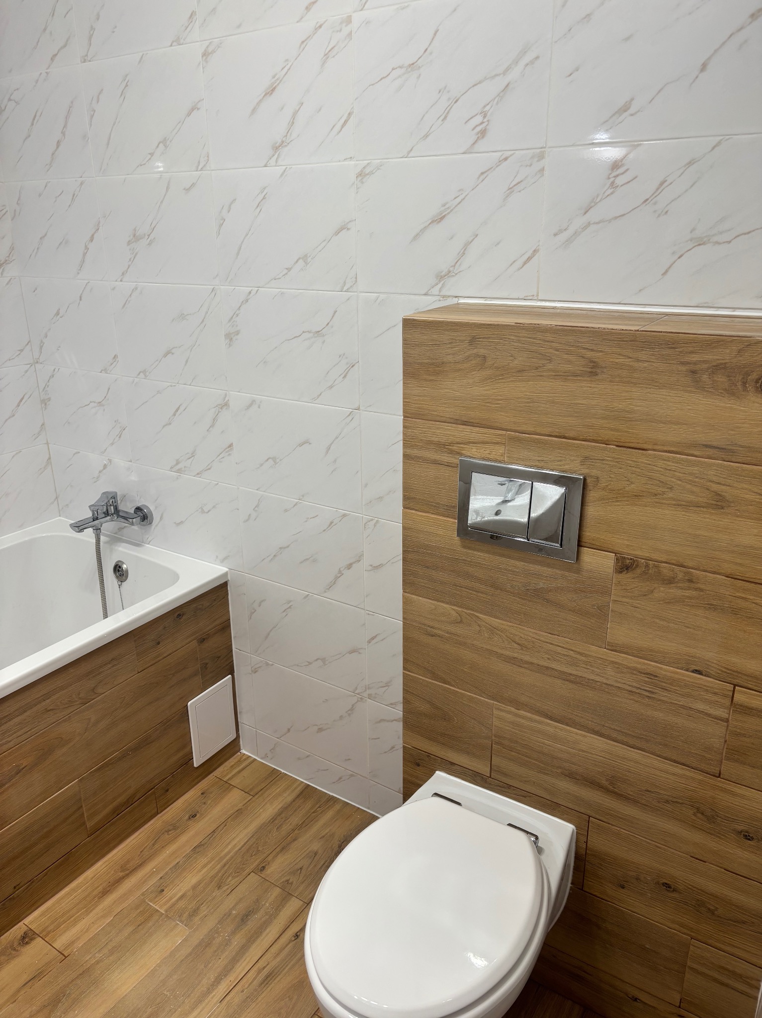 Nowoczesna łazienka z białymi płytkami imitującymi marmur na ścianach, drewnopodobnymi panelami na zabudowie wanny i stelażu WC, oraz podłogą w tym samym stylu.