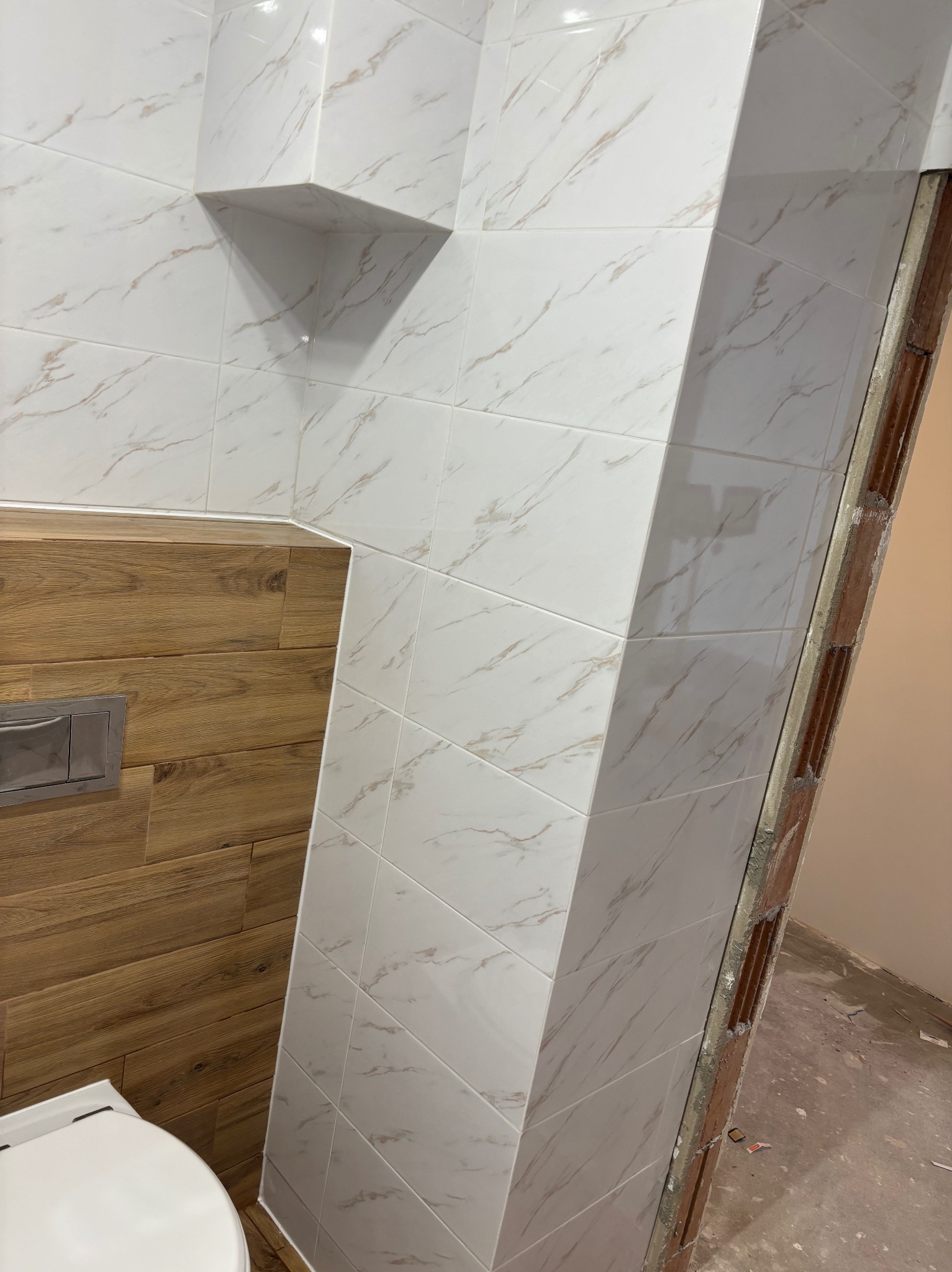 Białe płytki imitujące marmur z wnęką, drewniana obudowa stelaża WC z przyciskiem spłukującym, widoczny fragment toalety, surowa krawędź ściany z pozostałościami zaprawy.